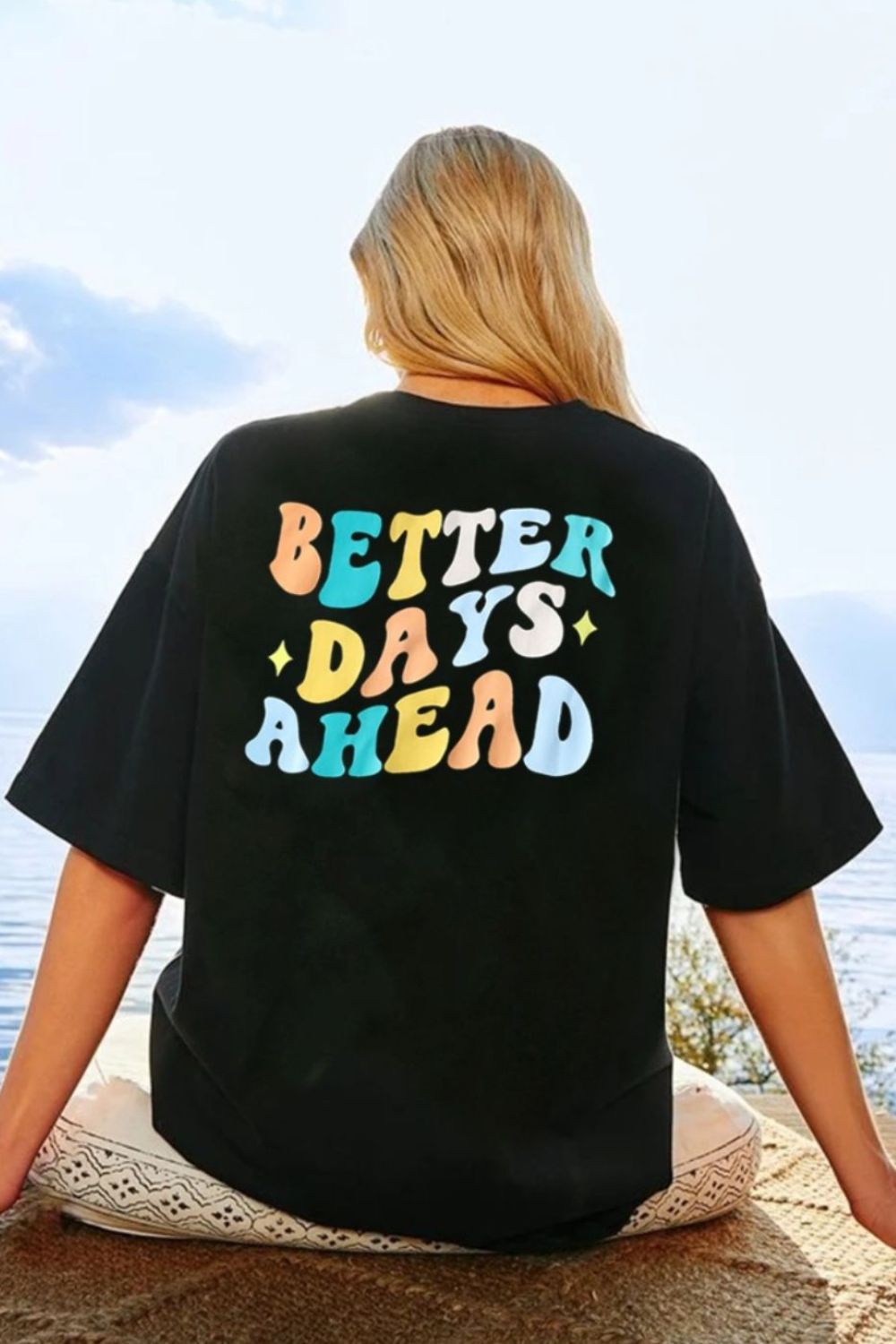 Better Days Ahead Graphic T-Shirt with colorful text design, worn by a person sitting outdoors by a lake.