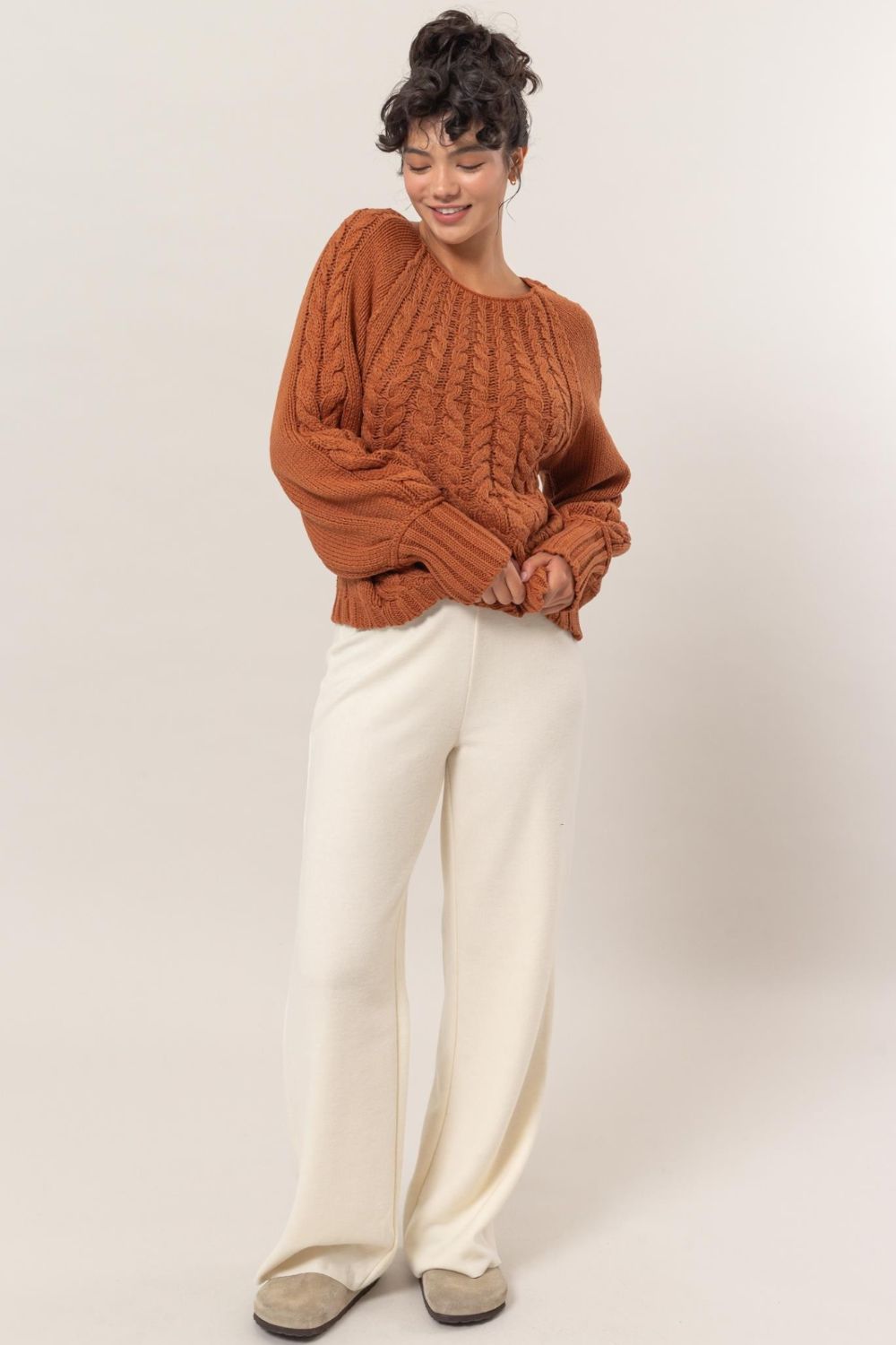 Cable knit round neck raglan sleeve sweater in orange, paired with white pants.