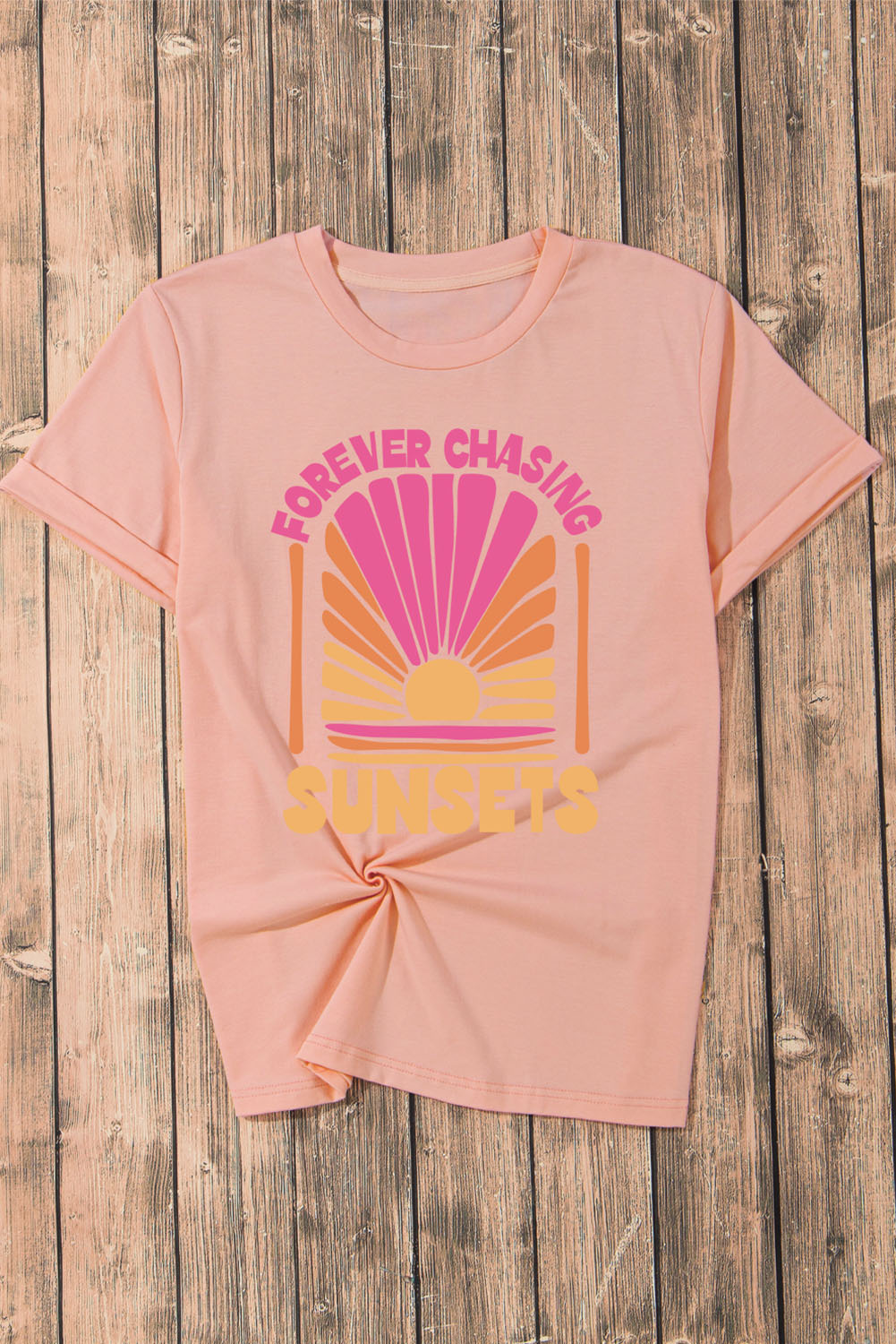 Forever Chasing Sunsets Graphic T-Shirt in pink featuring sunset design on wooden background.