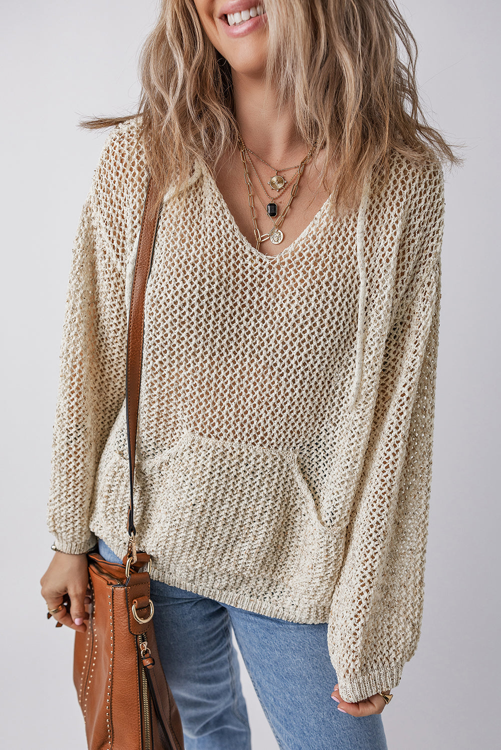 Openwork dropped shoulder hooded knit top with cutout design in beige, paired with jeans and a brown leather bag.