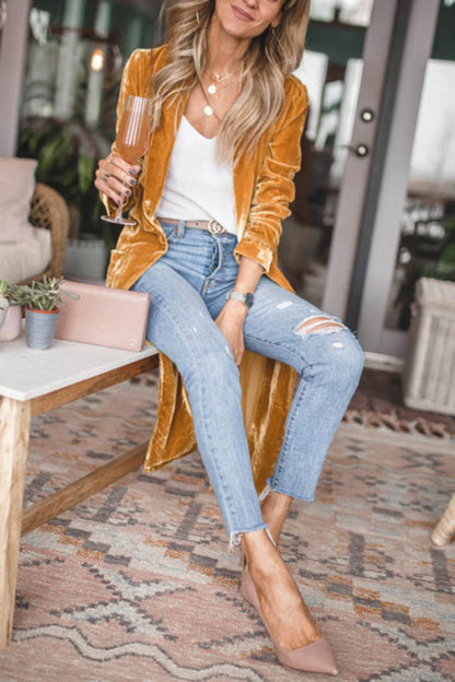 Woman wearing an open front long sleeve coat, paired with jeans and a white top, seated casually while holding a drink.