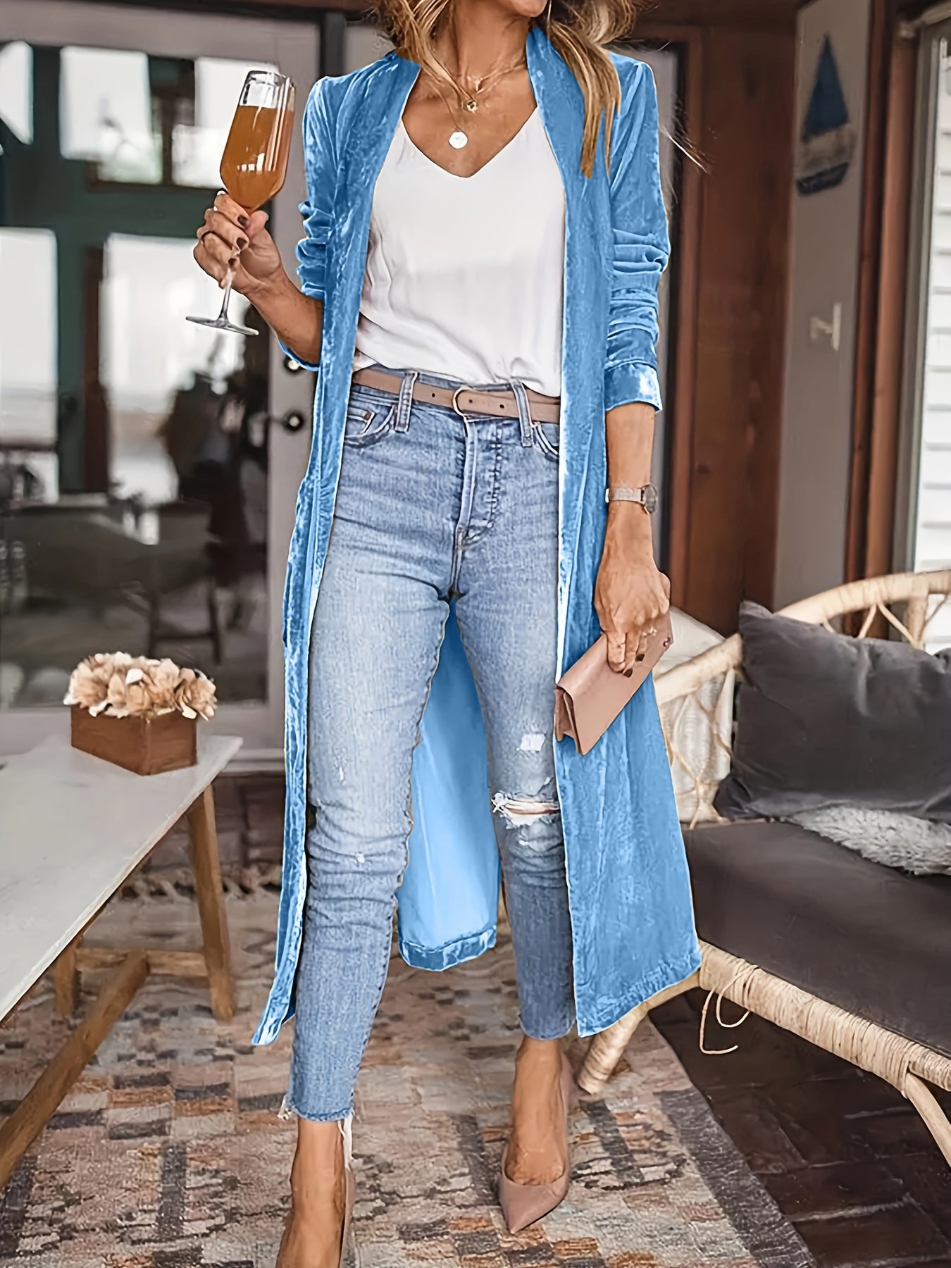 Woman wearing blue open-front long sleeve coat with jeans, holding a drink indoors.